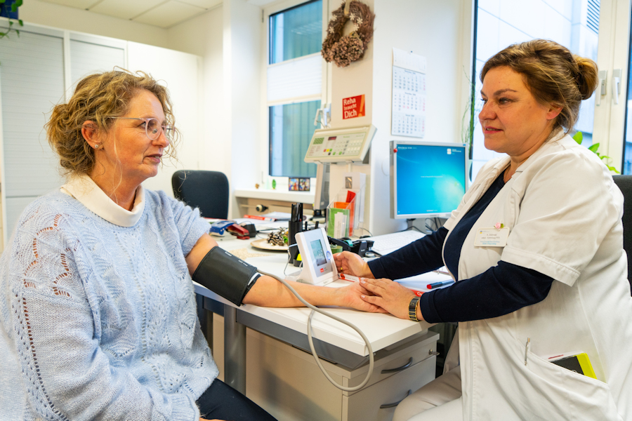 Gesundheits- und Krankenpflegerin misst mit beruhigeder Geste Blutdruck bei einer Rehabilitandin 